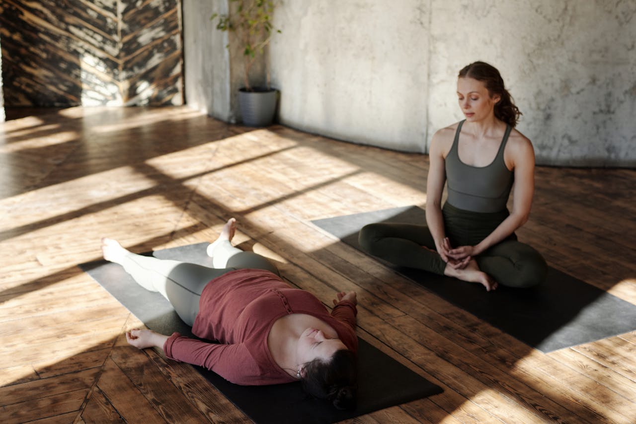 gallery-1 Two women practicing yoga indoors with sunlight streaming in, highlighting focus and relaxation.